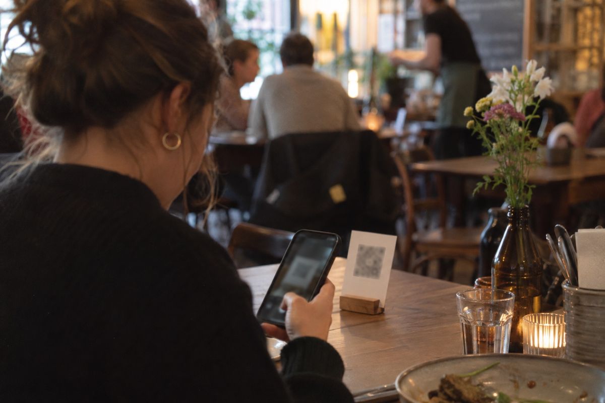 Ein Gast richtet am Holztisch eines Lokals das Smartphone auf den QR-Code-Aufsteller, um die digitale Speisekarte zu öffnen, daneben stehen Wasserglas, Windlicht und Blumen, im Hintergrund weitere Gäste und eine Servicekraft